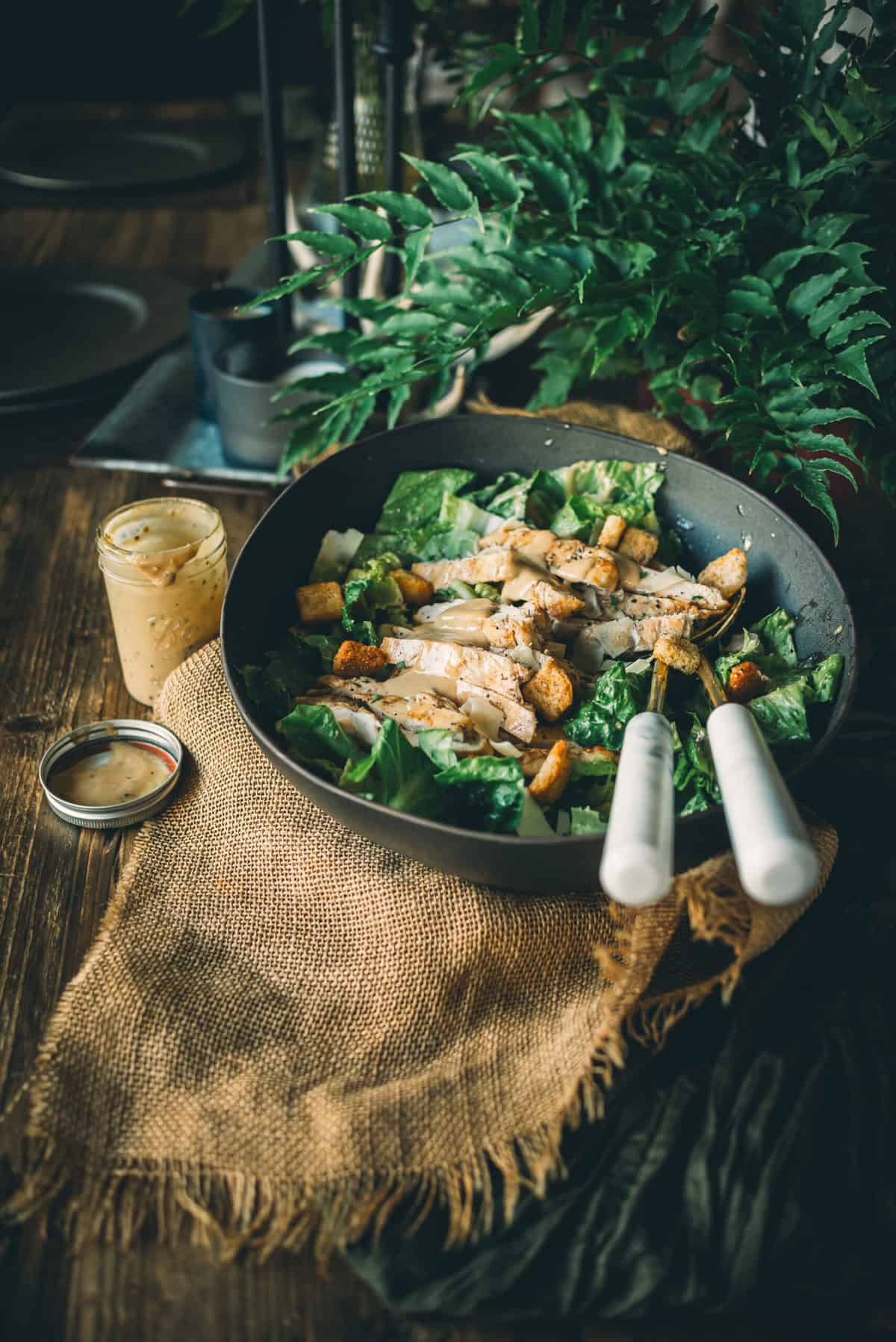 A bowl of Caesar salad with chicken, croutons, and parmesan cheese, placed on a wooden table with a small jar of dressing nearby and green plants in the background.