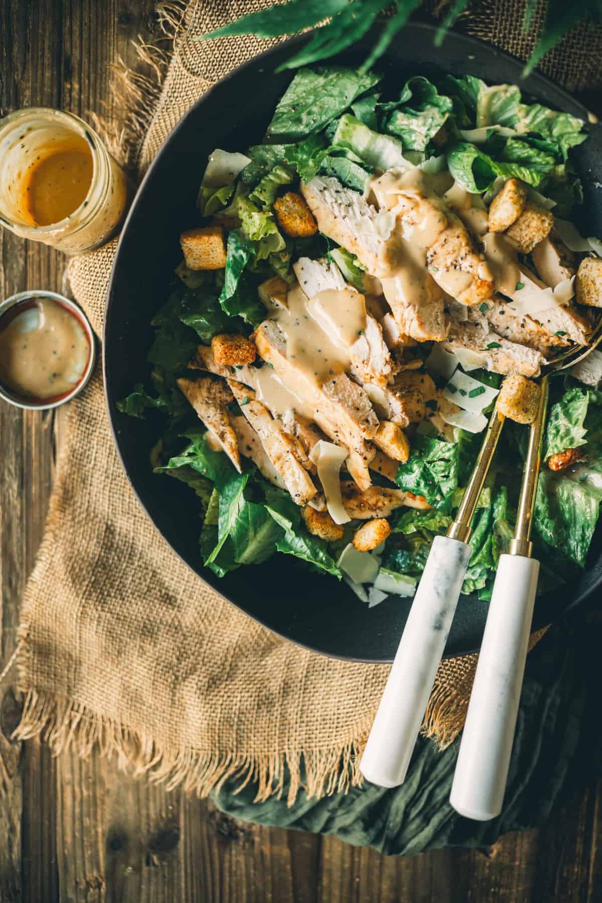 A bowl of Caesar salad with grilled chicken, croutons, and Caesar dressing, placed on a wooden table with a jar of extra dressing nearby.