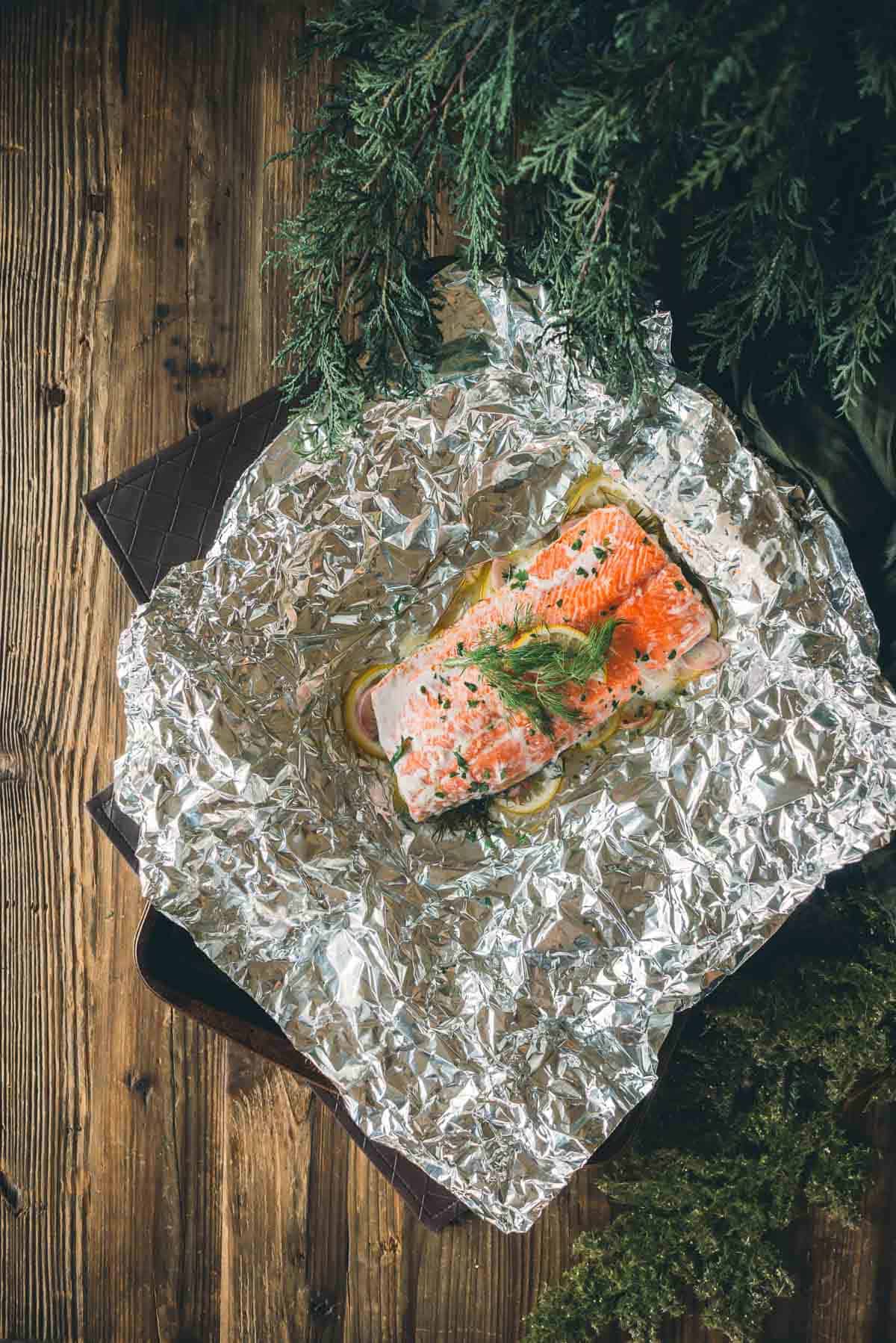 A cooked salmon fillet garnished with herbs sits on sliced lemons wrapped in aluminum foil, placed on a wooden surface with greenery in the background.