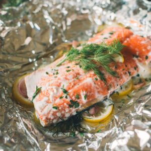 A piece of salmon topped with fresh herbs and lemon slices rests on a sheet of aluminum foil, ready for cooking.