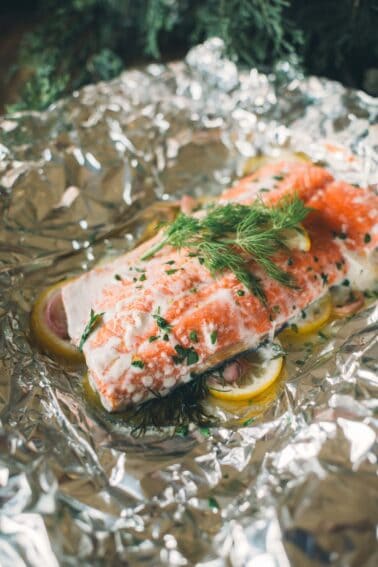 A piece of salmon topped with fresh herbs and lemon slices rests on a sheet of aluminum foil, ready for cooking.