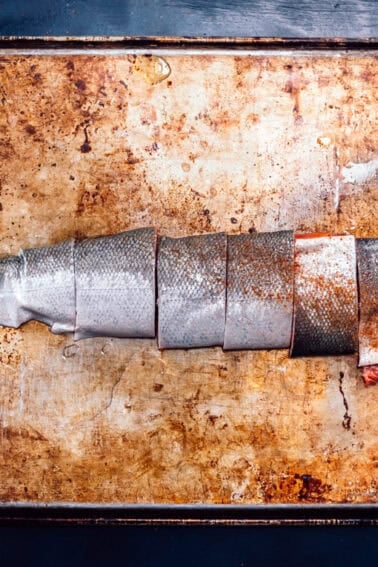A salmon fillet, with skin on, cut into four pieces, placed on a well-used, stained metal baking sheetโready to be transformed using the best wood for smoking salmon.