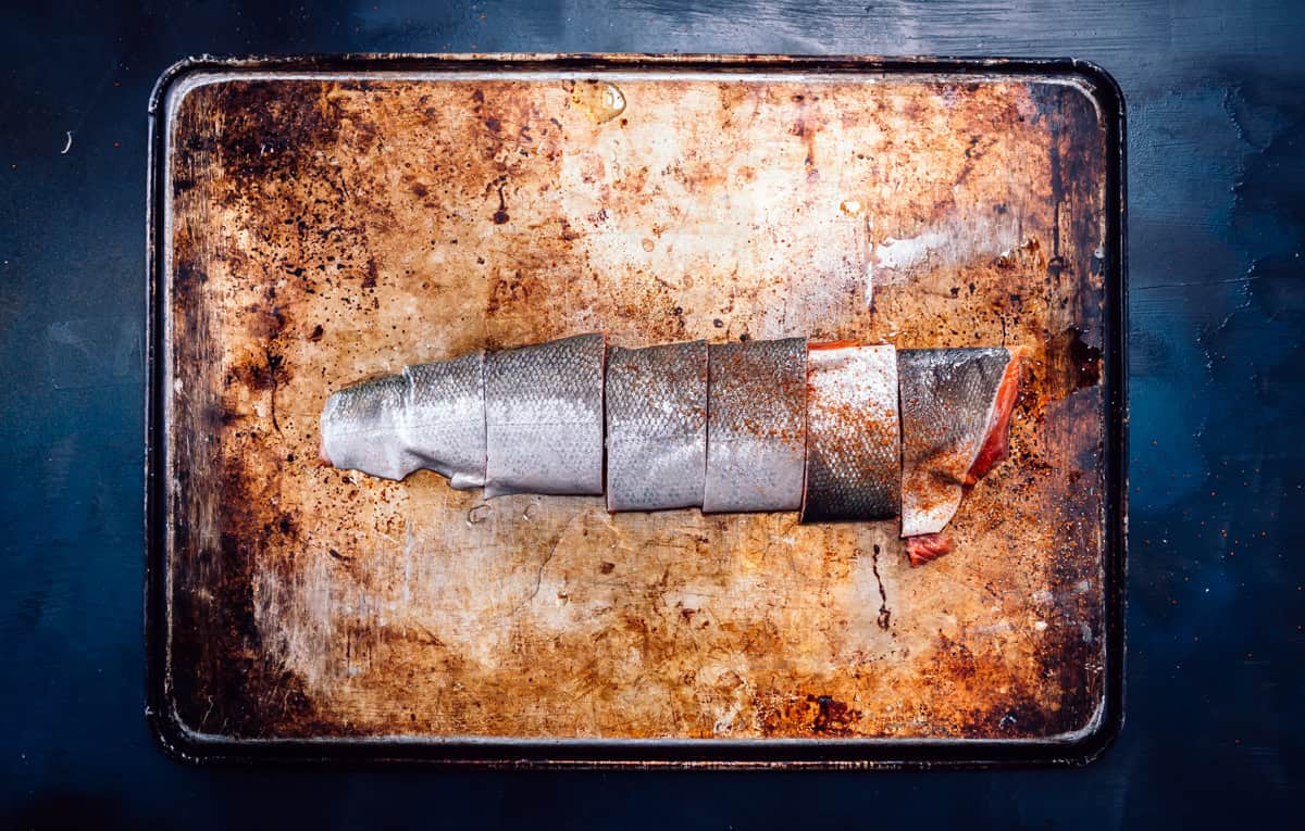 A salmon fillet, with skin on, cut into four pieces, placed on a well-used, stained metal baking sheet—ready to be transformed using the best wood for smoking salmon.