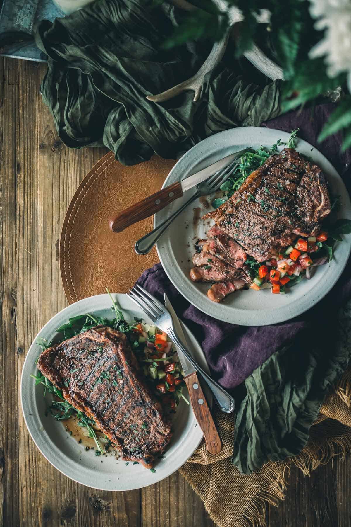 Two plates of grilled steak served on a bed of greens with a side of diced vegetables.