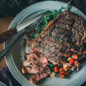 A plate with a cooked KC strip steak partially sliced, garnished with herbs, atop a bed of salad featuring diced red peppers.