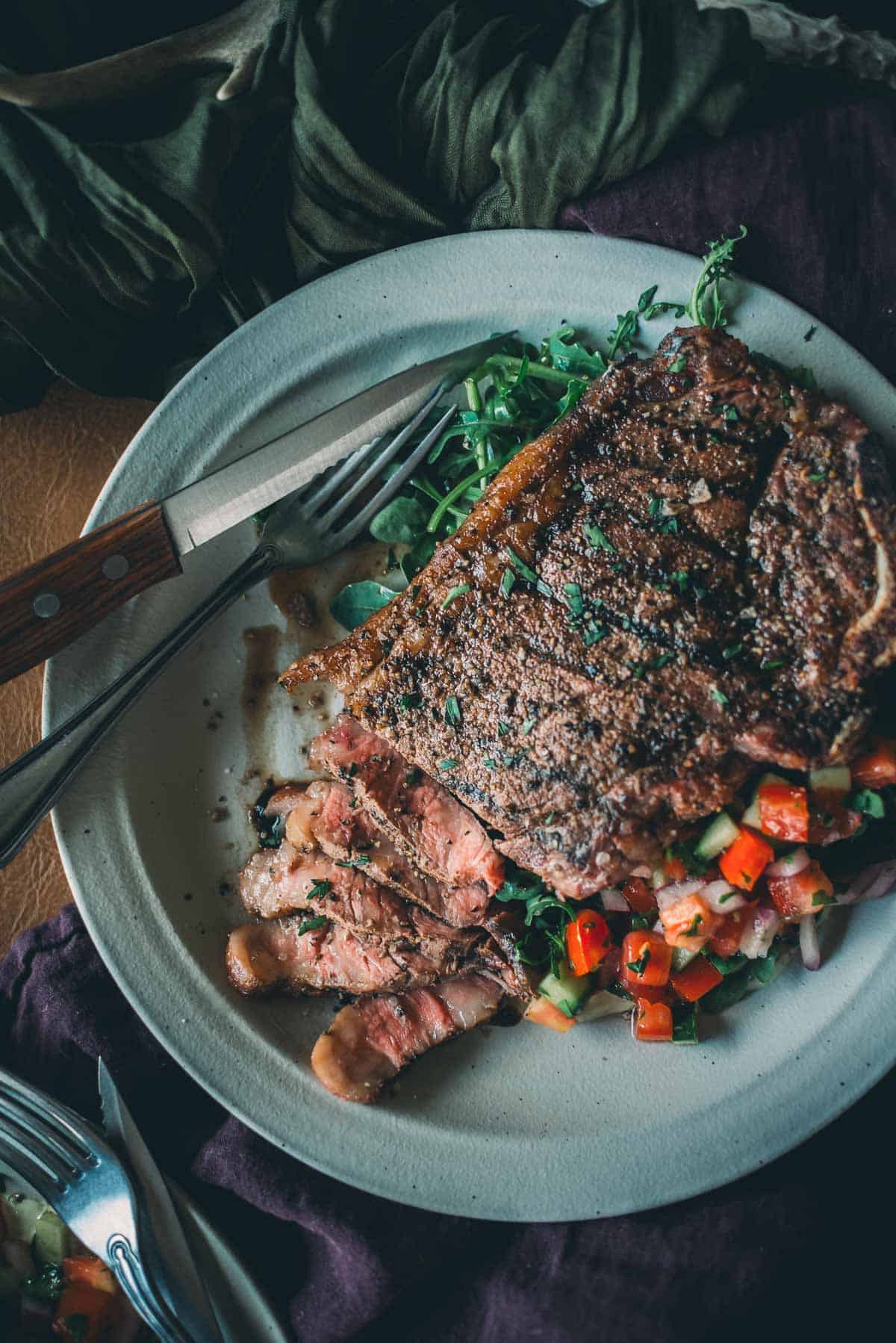 A plate with a cooked KC strip steak partially sliced, garnished with herbs, atop a bed of salad featuring diced red peppers.