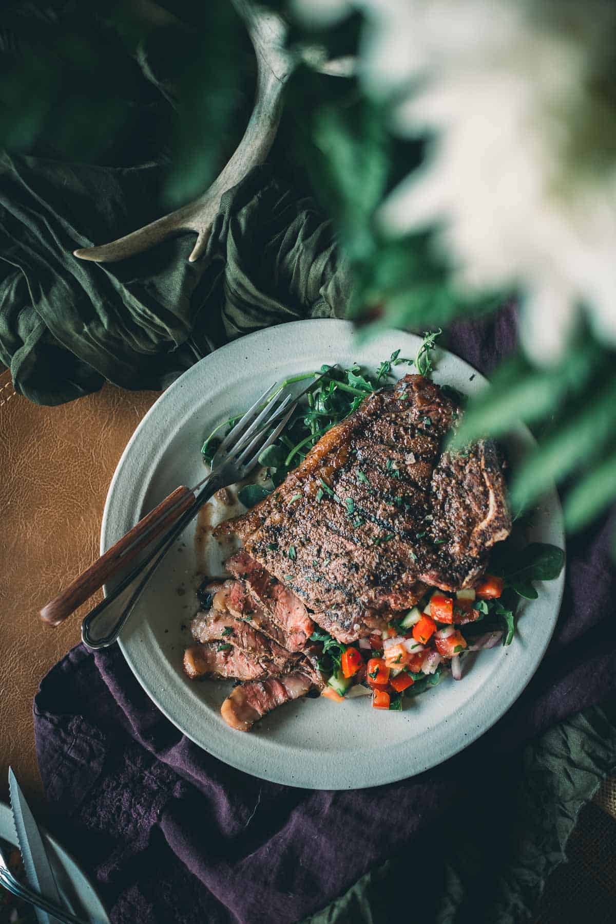 A plate of grilled strip steak partially sliced, garnished with chopped vegetables and greens, with a fork and knife placed beside it on a cloth-covered table.