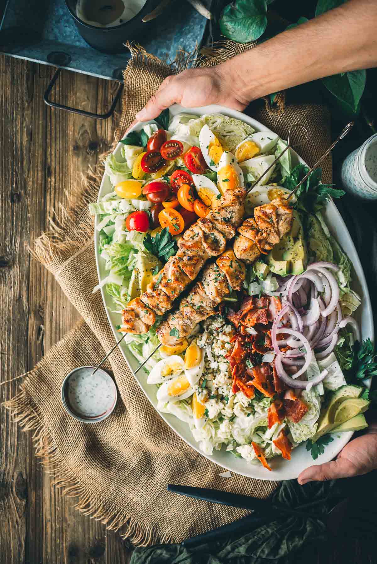 A platter of fresh salad topped with grilled chicken skewers, cherry tomatoes, hard-boiled eggs, red onions, bacon, and avocado slices, garnished with a side of dressing.