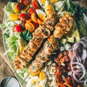 Appetizing cobb salad topped with grilled chicken skewers, cherry tomatoes, avocado, bacon, hard-boiled eggs, red onions, blue cheese, and lettuce.