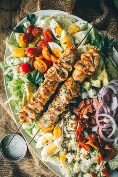 Appetizing cobb salad topped with grilled chicken skewers, cherry tomatoes, avocado, bacon, hard-boiled eggs, red onions, blue cheese, and lettuce.
