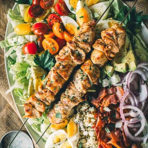 Appetizing cobb salad topped with grilled chicken skewers, cherry tomatoes, avocado, bacon, hard-boiled eggs, red onions, blue cheese, and lettuce.