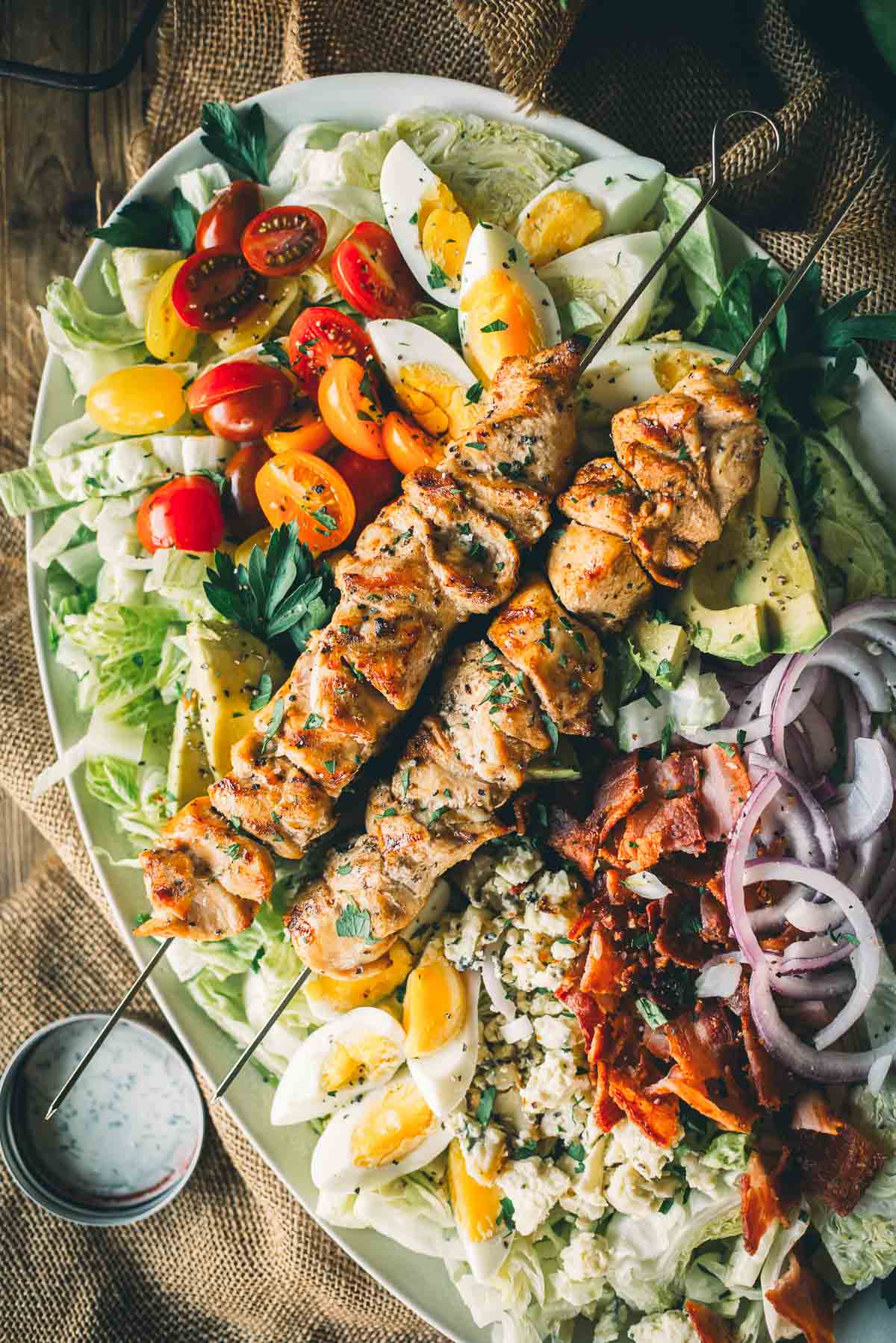 Appetizing cobb salad topped with grilled chicken skewers, cherry tomatoes, avocado, bacon, hard-boiled eggs, red onions, blue cheese, and lettuce.