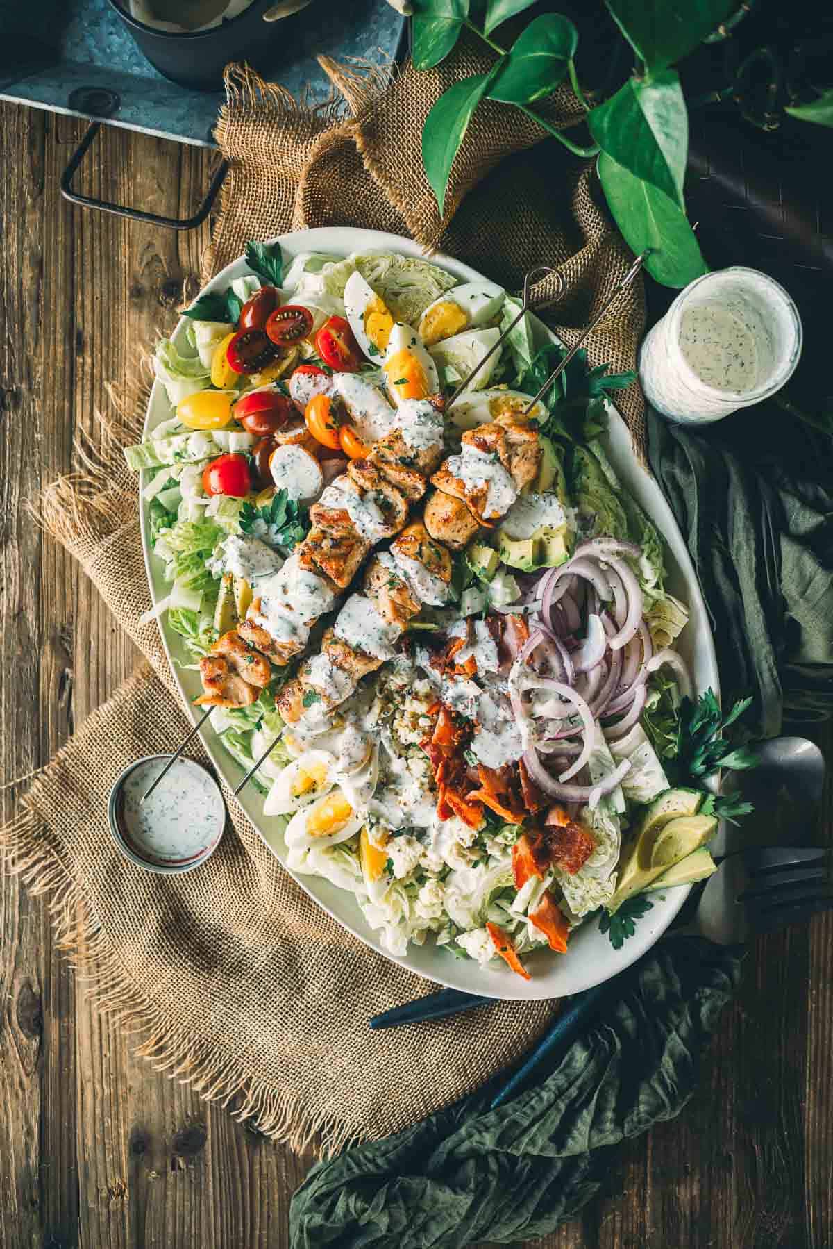 A platter of grilled chicken cobb salad with mixed vegetables, boiled eggs, and chicken skewers, drizzled with creamy dressing, on a wooden table.