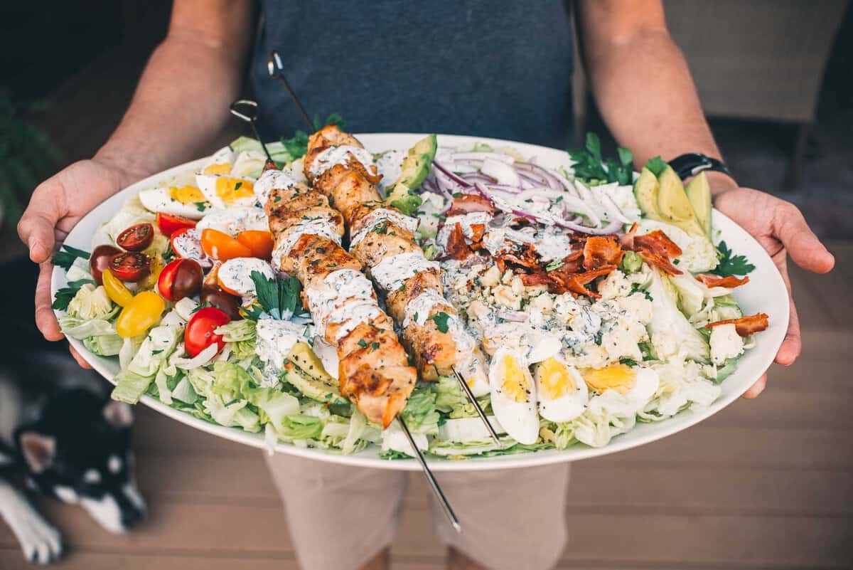 Person holding a large plate of salad featuring grilled chicken skewers, cherry tomatoes, hard-boiled eggs, red onions, bacon bits, and avocado slices on a bed of lettuce.