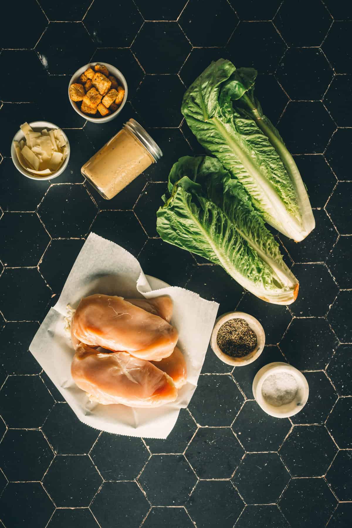 Ingredients for Caesar salad arranged on a black tile surface, including chicken breasts, romaine lettuce, Caesar dressing, croutons, Parmesan cheese, salt, and pepper.