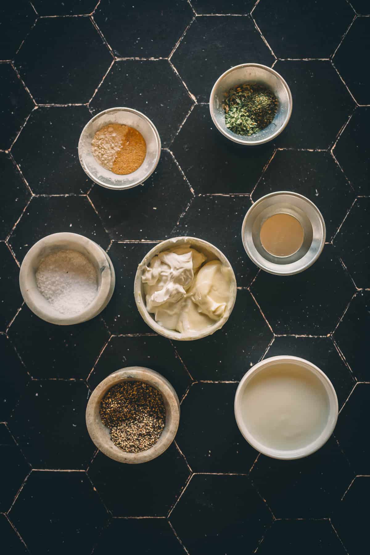 Seven bowls containing various ingredients for creamy caesar dressing are arranged on a black surface.