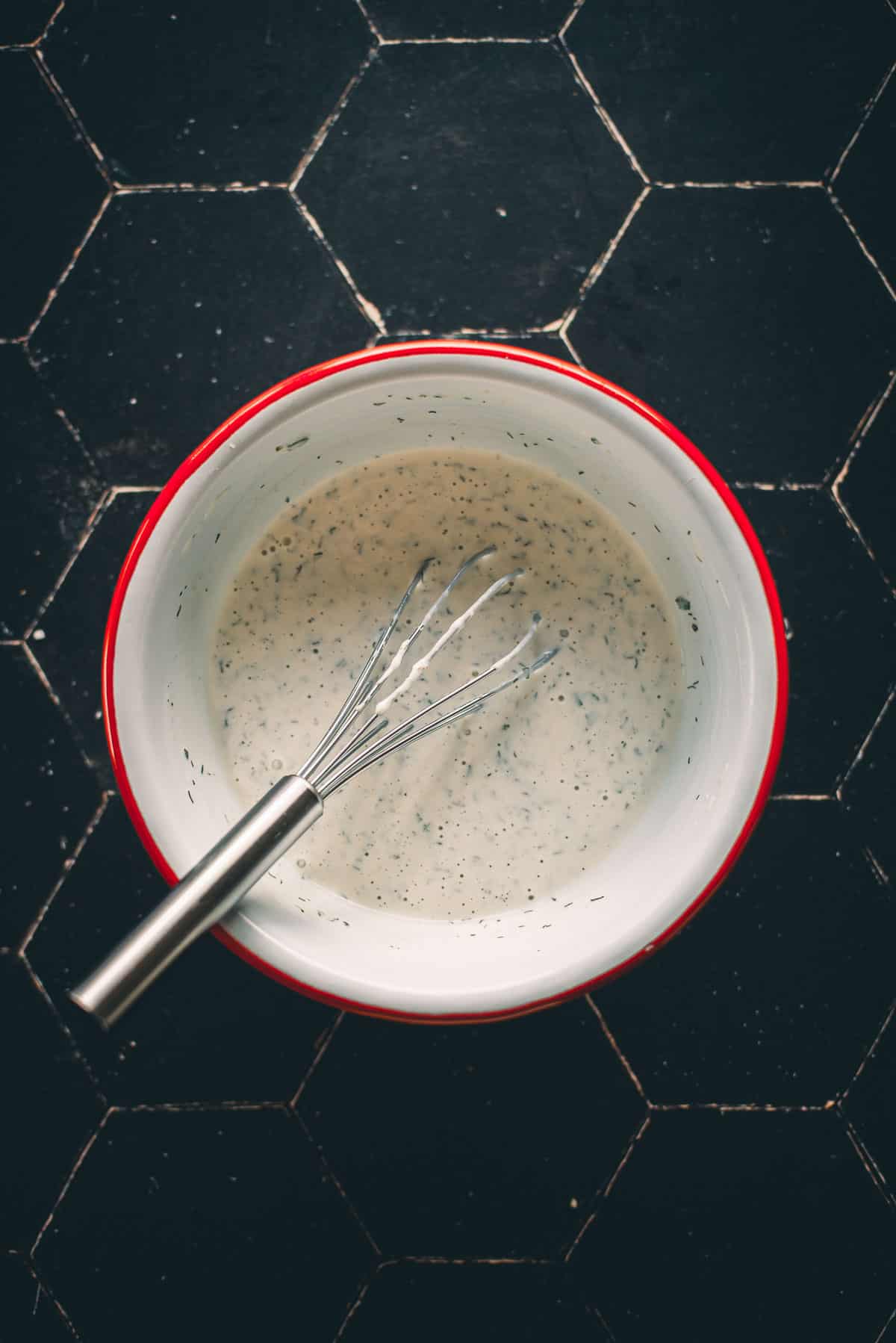 A metal whisk in a red and white bowl filled with a creamy dressing, placed on a black surface.