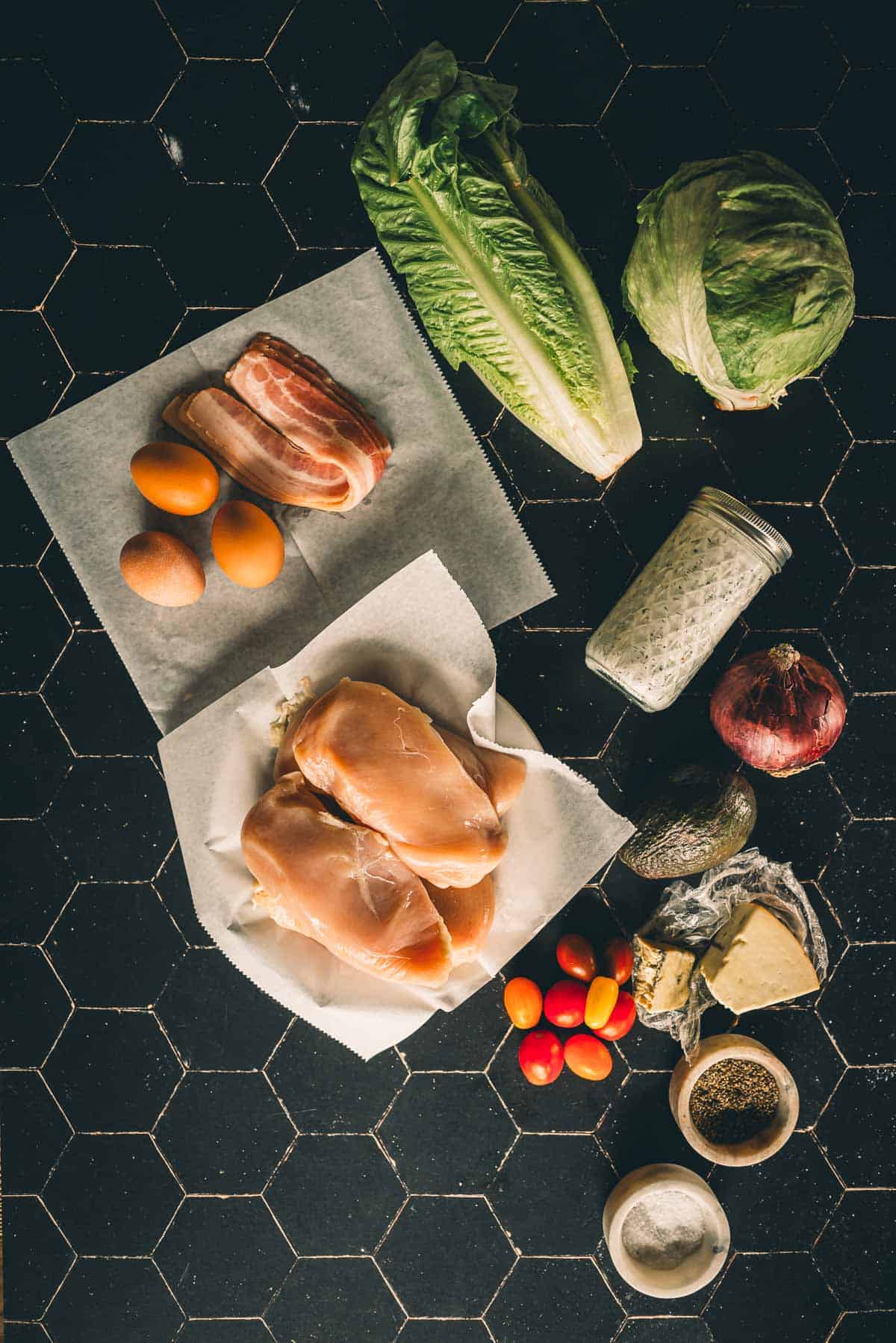 Various ingredients on a black tile surface, including chicken breasts, eggs, bacon, lettuce, cabbage, spices, tomatoes, onion, avocado, cheese, and a jar of dressing.