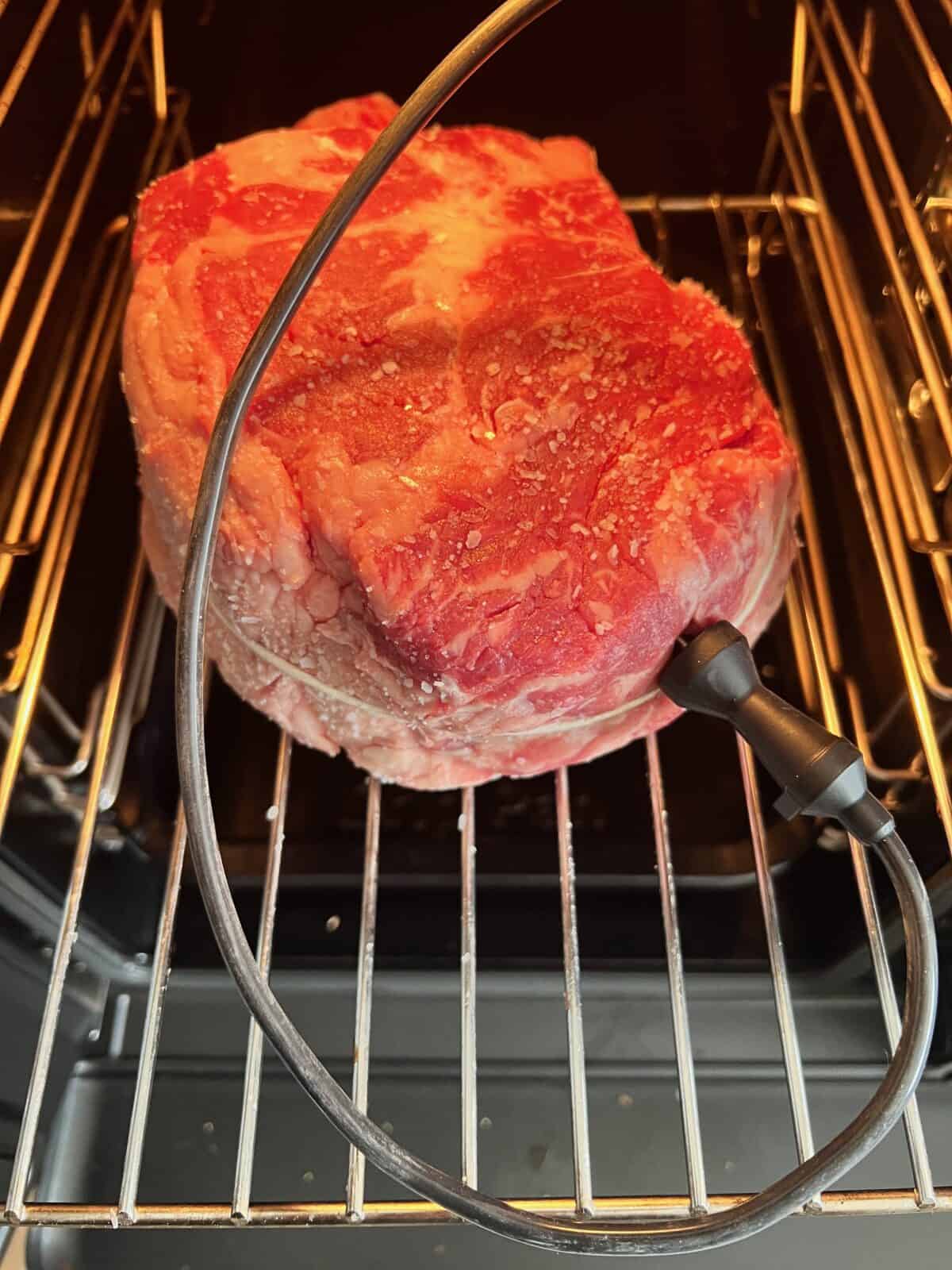 A raw cowboy steak with salt is placed in a smoker on a metal rack, with a meat thermometer probe inserted into it.