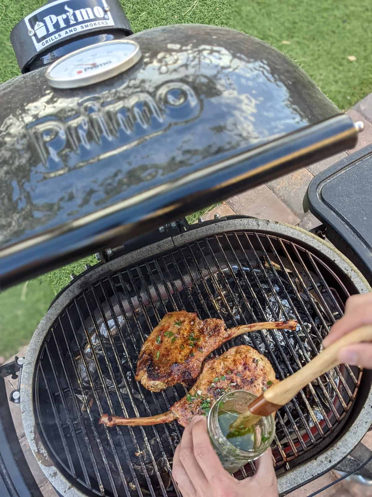 A person is expertly applying sauce to two grilled tomahawk pork chops cooking on a Primo grill, demonstrating how to use a kamado grill to achieve perfect flavor and sear.