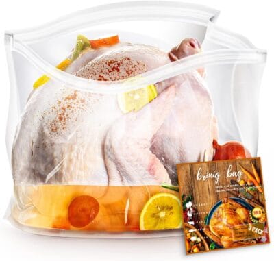 Image of a raw turkey and vegetables in a brining bag with a packet of brine seasoning. The bag has lemon slices, carrots, and other vegetables inside with the turkey.