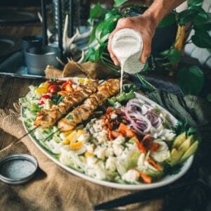 A person pours creamy dressing over a large platter of mixed salad with grilled chicken skewers, vegetables, and greens.