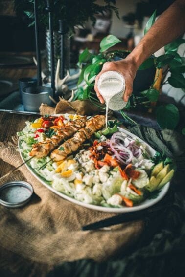 A person pours creamy dressing over a large platter of mixed salad with grilled chicken skewers, vegetables, and greens.