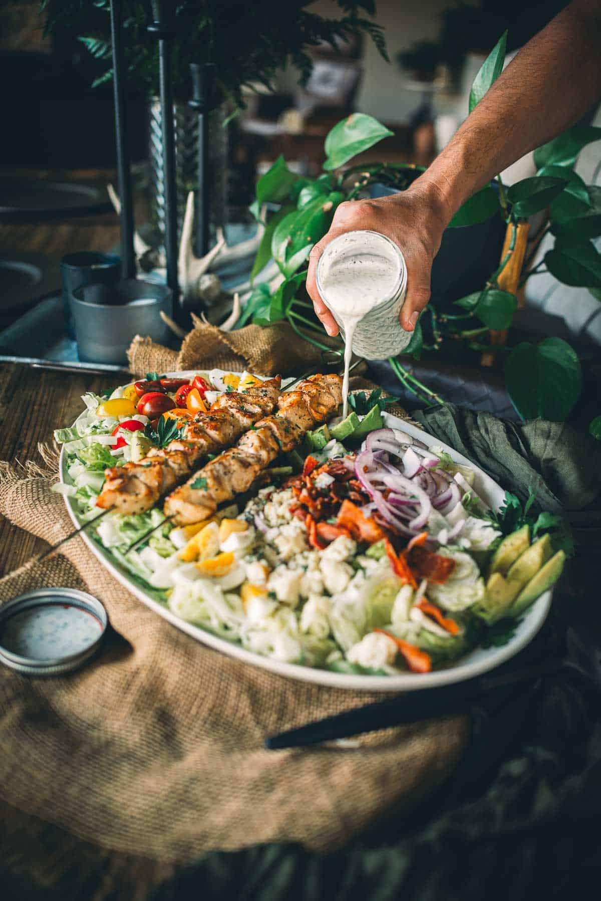 A person pours creamy dressing over a large platter of mixed salad with grilled chicken skewers, vegetables, and greens.