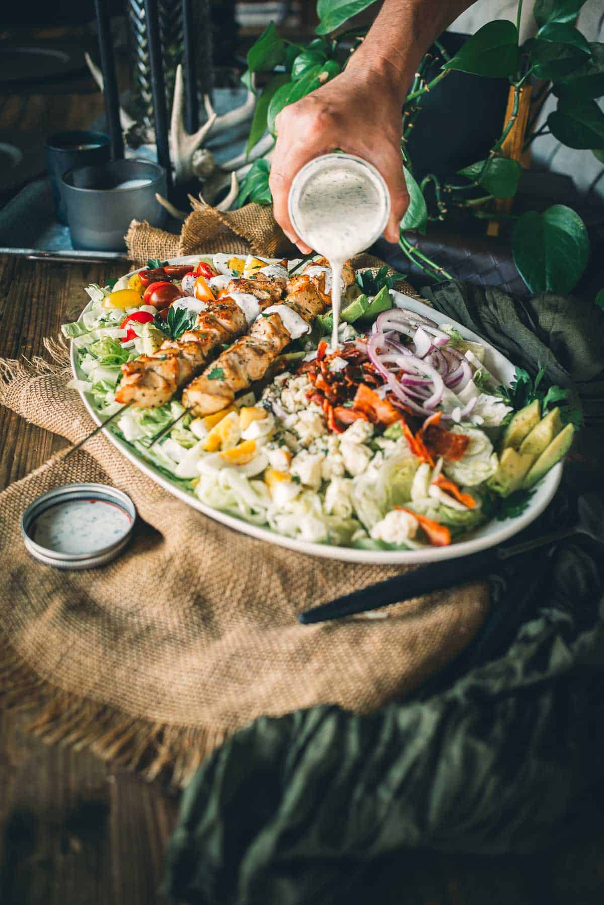 A person is pouring ranch dressing over a large, colorful salad that includes grilled chicken, red onions, bacon, lettuce, and tomatoes, set on a rustic wooden table.