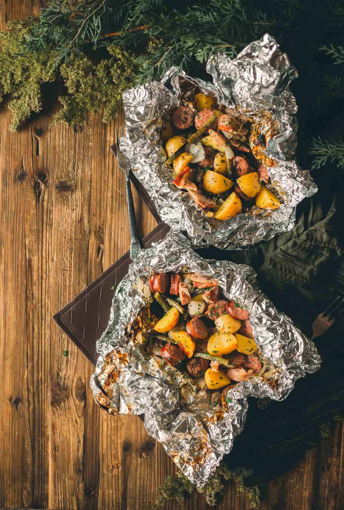 Two foil packets on a wooden surface contain diced potatoes, sliced sausages, and vegetables, garnished with herbs. 