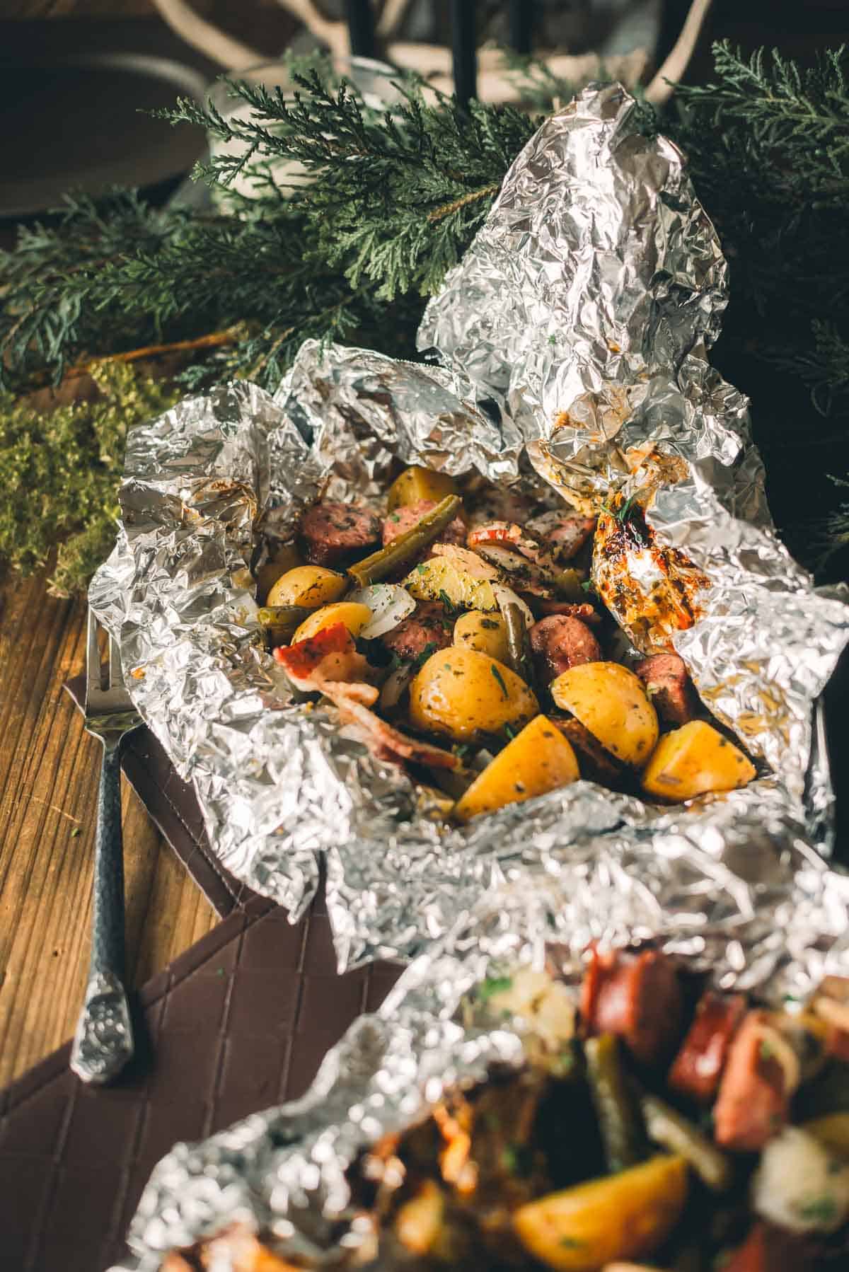 Foil packets filled with chopped potatoes, sausages, and vegetables are placed on a wooden table next to a fork and some greenery.