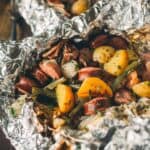 Close-up of a meal wrapped in aluminum foil containing sliced sausage, chunks of potato, green beans, onions, and herbs.