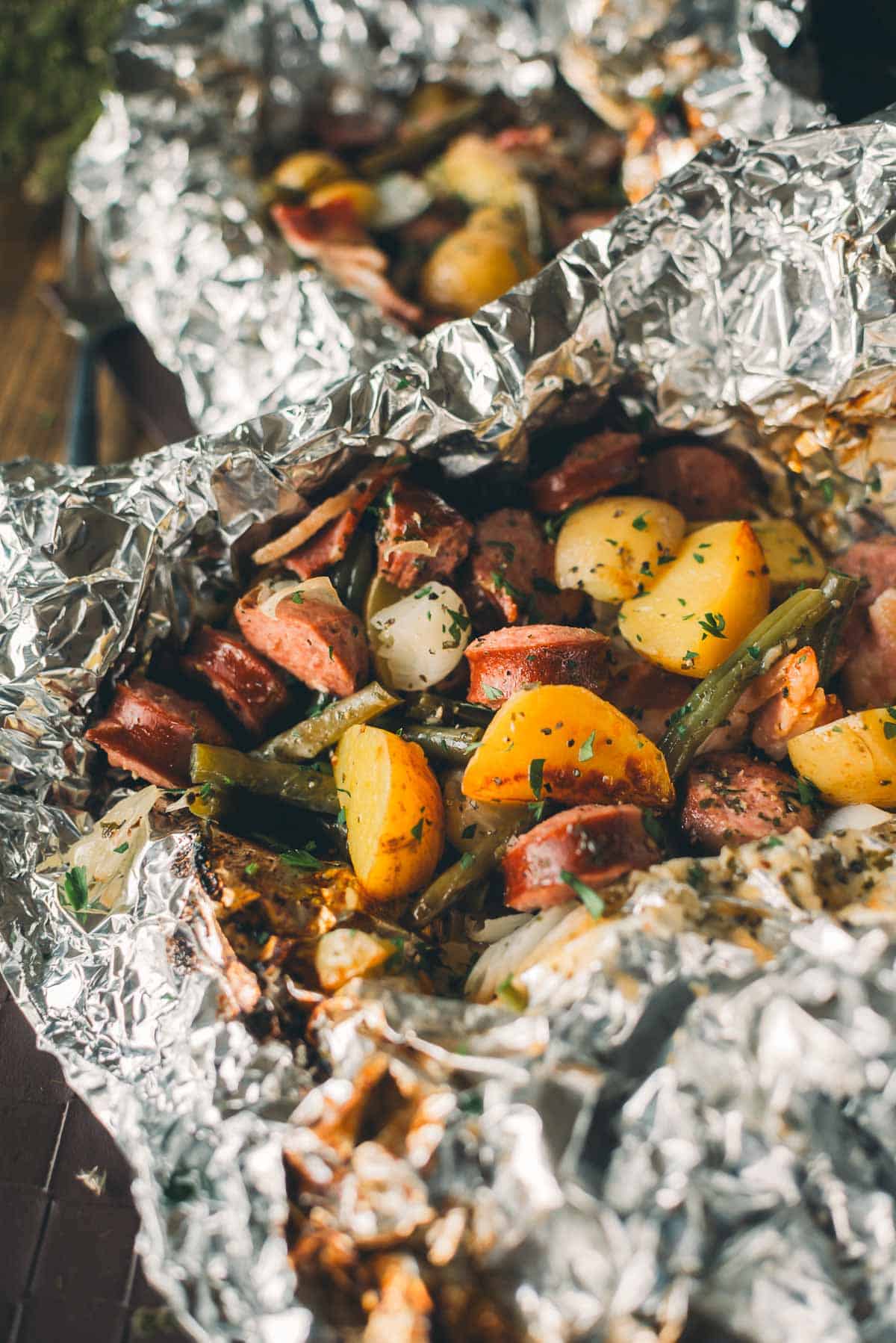 Close-up of a meal wrapped in aluminum foil containing sliced sausage, chunks of potato, green beans, onions, and herbs.