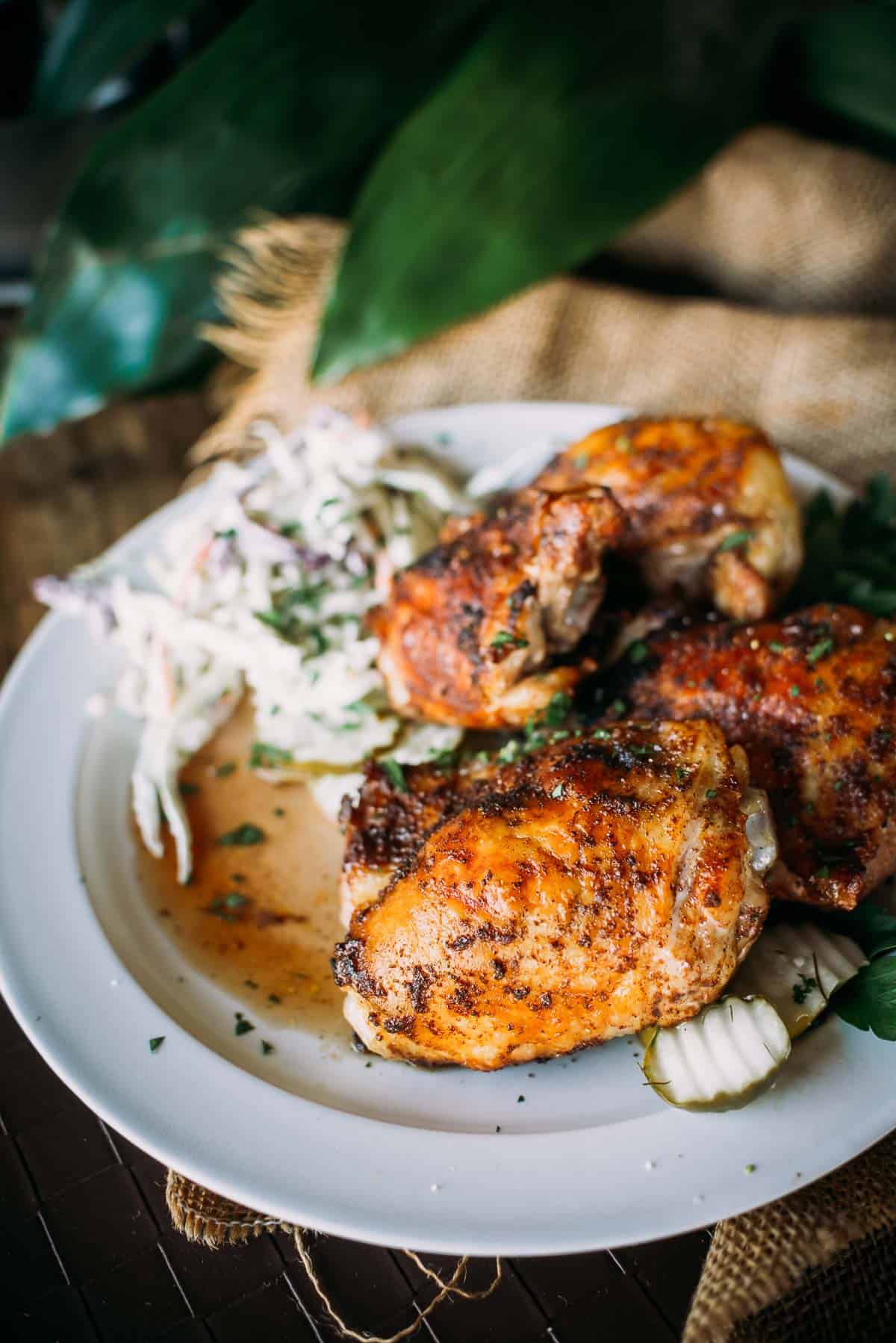 A white plate with grilled chicken thighs seasoned with herbs and served with a side of coleslaw.