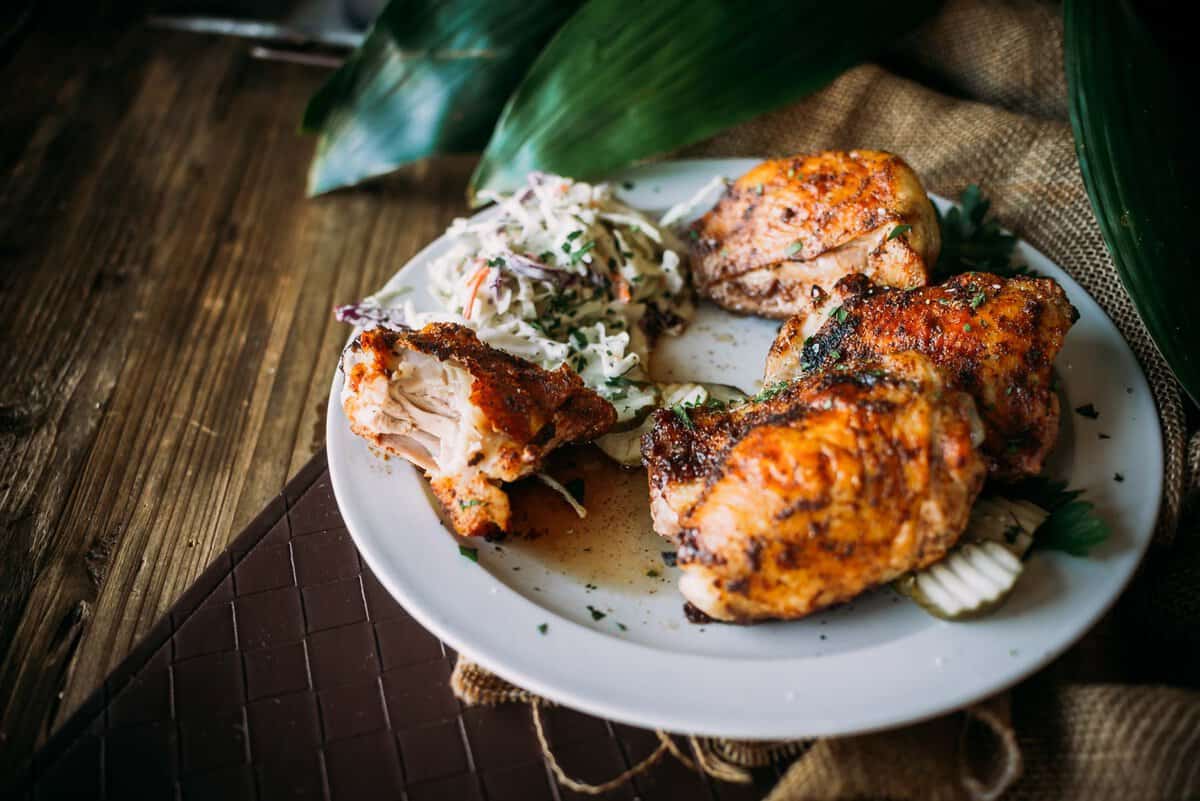 A plate of seasoned grilled chicken pieces is served with a side of coleslaw on a wooden table with leafy greens in the background.