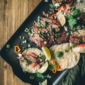 A tray with grilled skirt steak tacos garnished with lime wedges, cilantro, green onions, crumbled cheese, and sliced red peppers on flour tortillas.