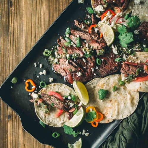 A tray with grilled skirt steak tacos garnished with lime wedges, cilantro, green onions, crumbled cheese, and sliced red peppers on flour tortillas.