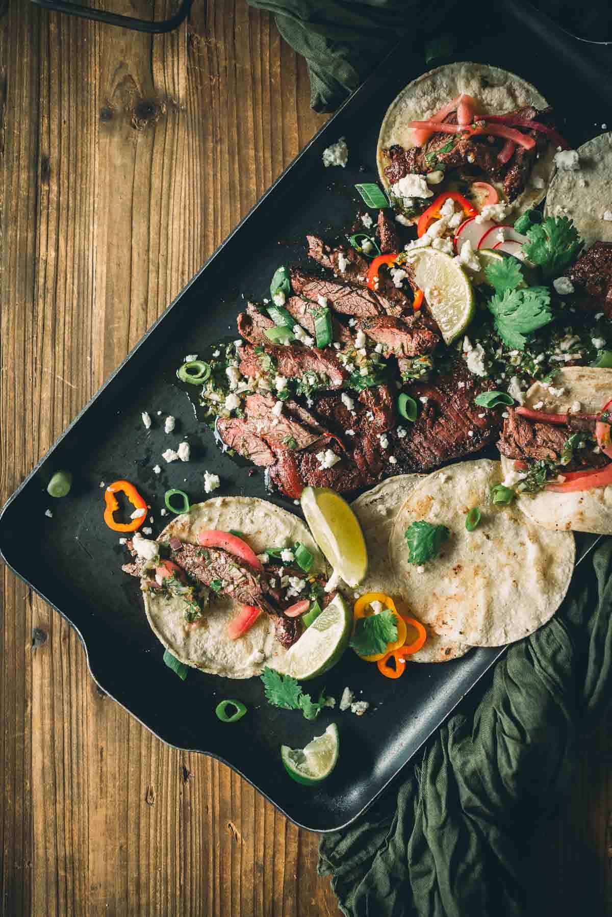 A tray with grilled skirt steak tacos garnished with lime wedges, cilantro, green onions, crumbled cheese, and sliced red peppers on flour tortillas.