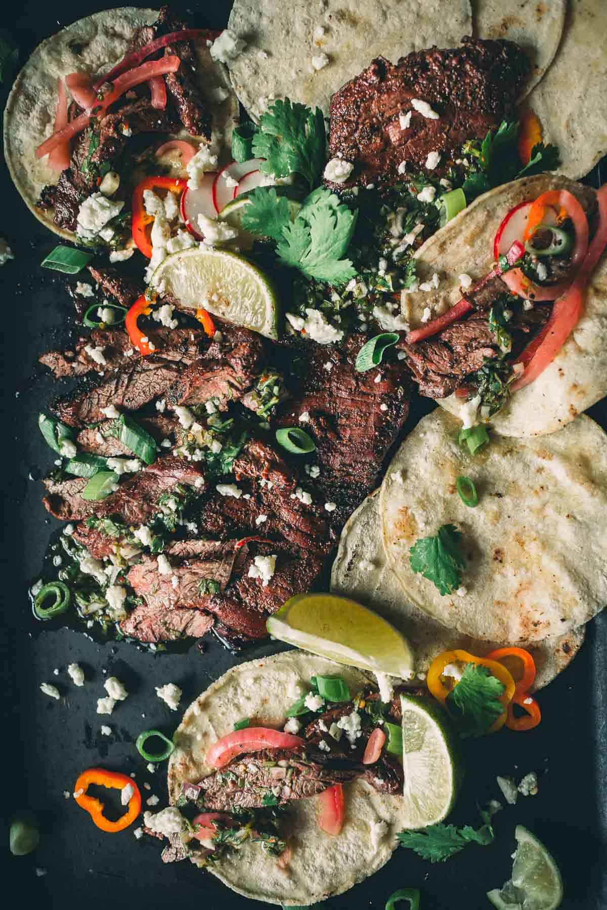 A platter of tacos featuring grilled slices of skirt steak, lime wedges, chopped cilantro, sliced red onions, and colorful bell pepper rings on a dark surface.