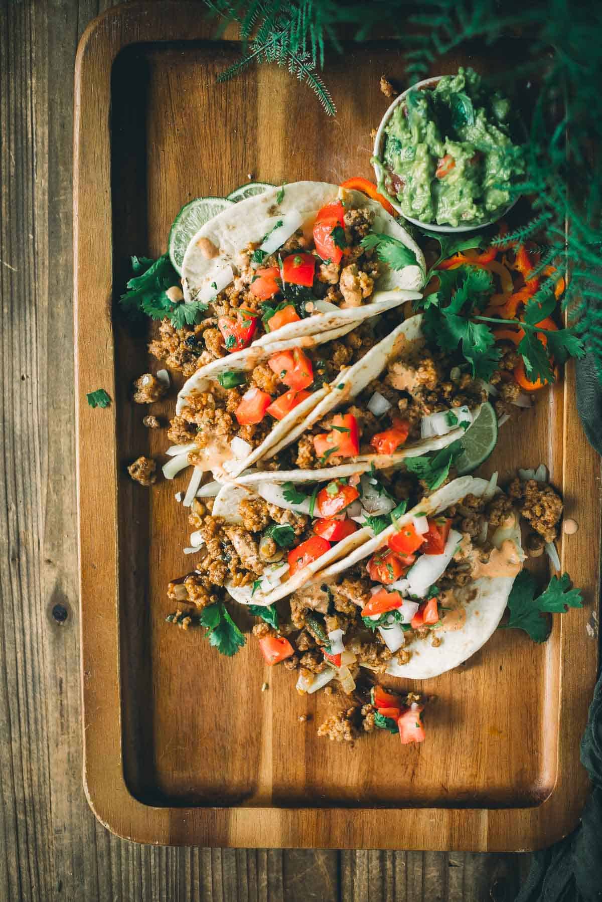 Wooden tray with three soft shell tacos filled with mixed vegetables, minced meat, and topped with chopped tomatoes, onions, and cilantro. A small bowl of guacamole is placed beside the tacos.