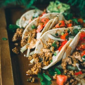 A wooden tray with four tacos filled with ground meat, chopped tomatoes, onions, and cilantro, garnished with avocado slices in the background.