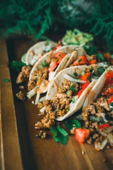 A wooden tray with four tacos filled with ground meat, chopped tomatoes, onions, and cilantro, garnished with avocado slices in the background.