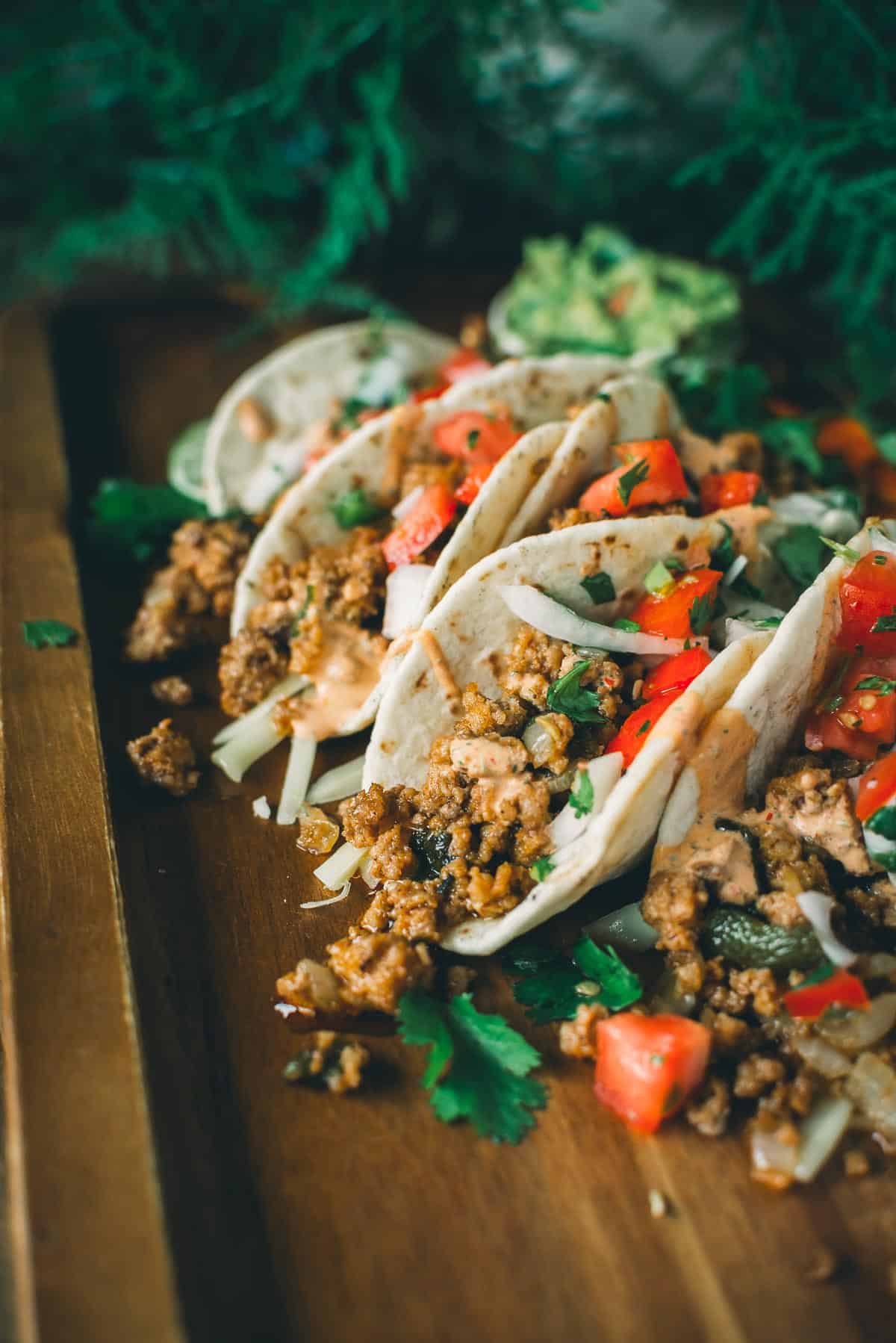 A wooden tray with four tacos filled with ground meat, chopped tomatoes, onions, and cilantro, garnished with avocado slices in the background.