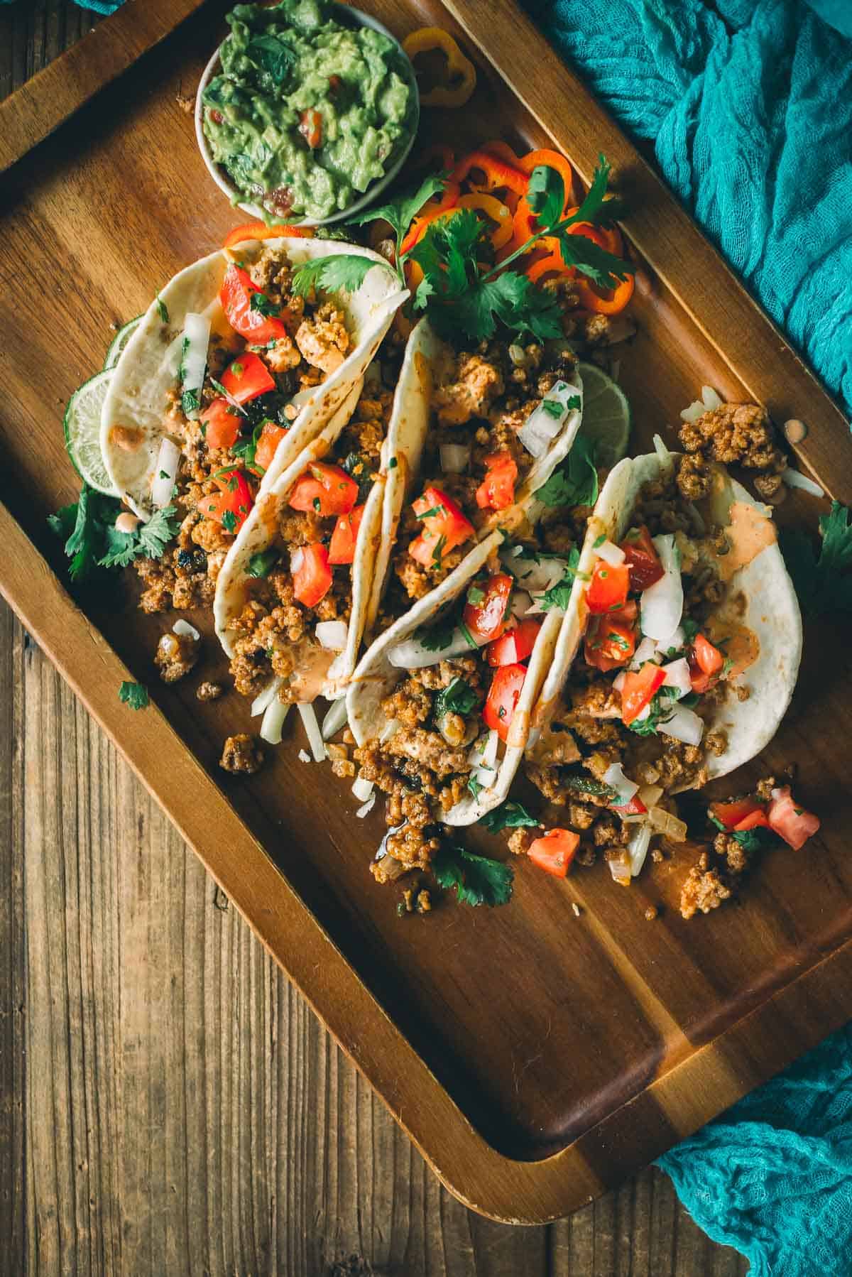 A wooden platter with four tacos filled with seasoned ground meat, diced tomatoes, onions, and cilantro. A small bowl of guacamole and sliced bell peppers are on the side.