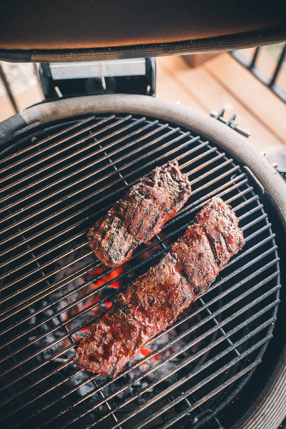 Two skirt steaks are being grilled on a round barbecue grill with visible hot coals beneath.