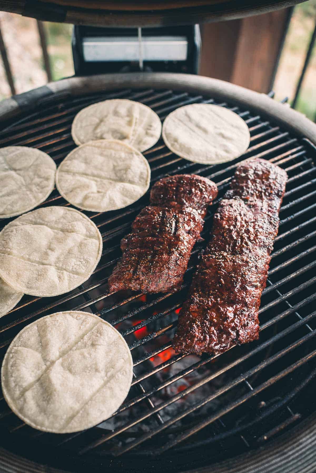 Two skirt steaks and several corn tortillas are cooking on a round outdoor grill.