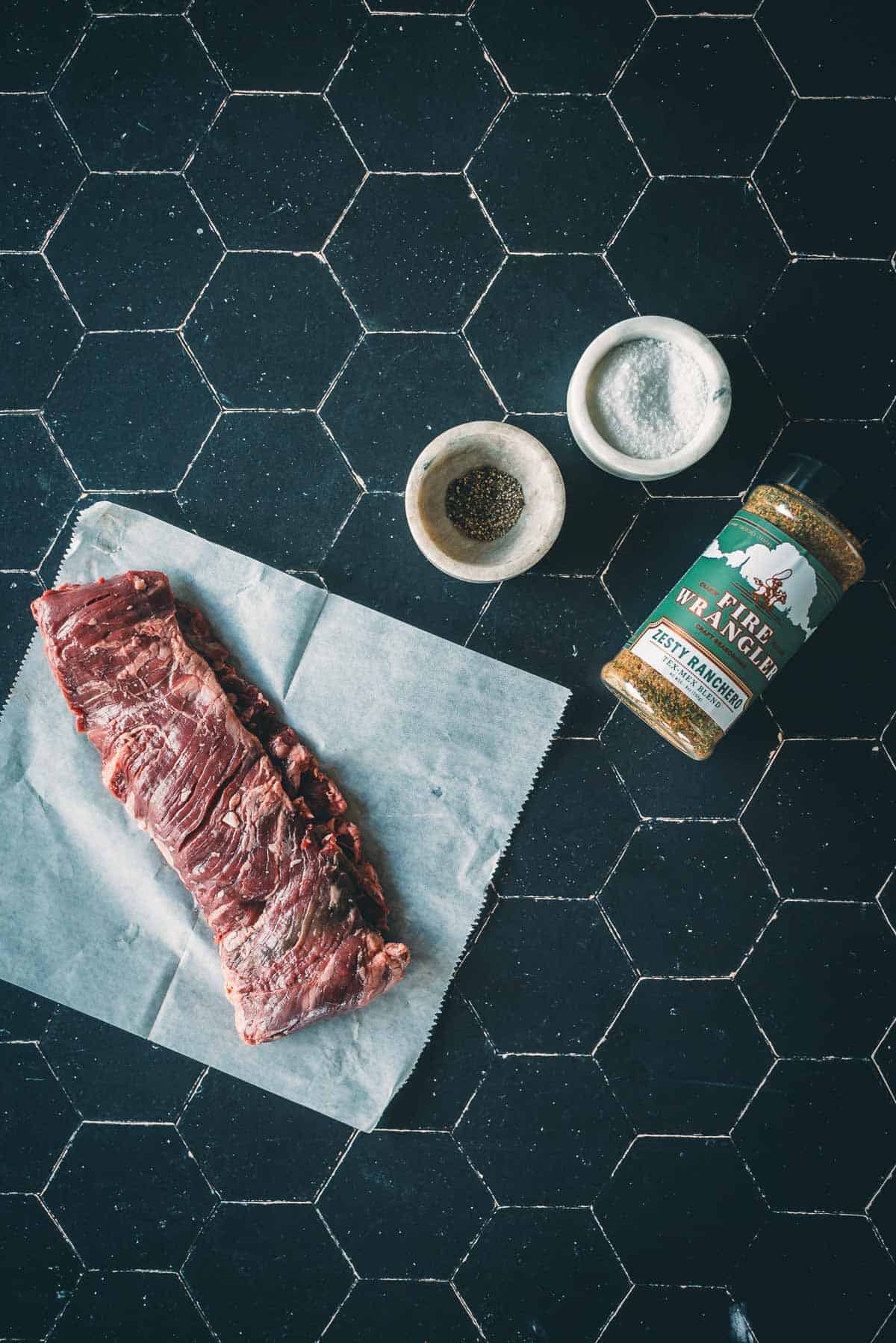 A piece of raw skirt steak on parchment paper next to bowls of salt and pepper, and a container of seasoning, all placed on a black tile surface.