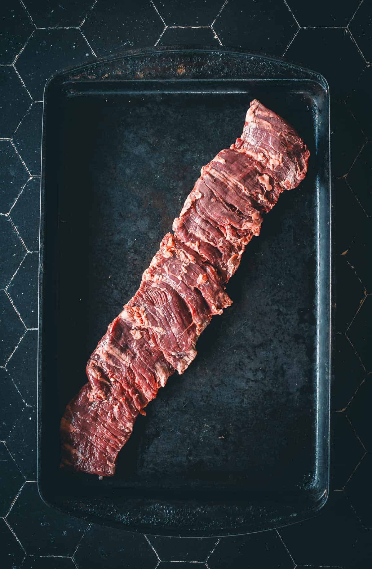 A raw skirt steak lies on a black baking sheet to show the unique grain of the cut.