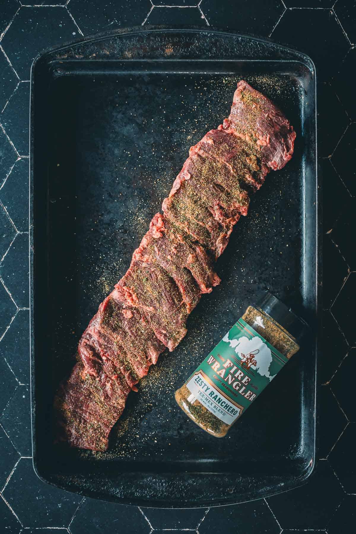 A seasoned strip of raw skirt steak lies on a black baking tray next to a container seasoning.