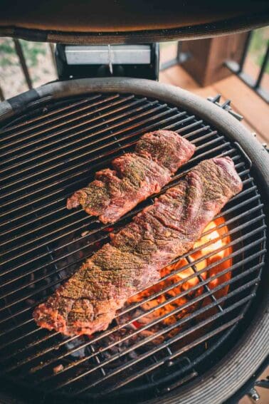 Two seasoned racks of ribs cooking on a grill with visible flames beneath.